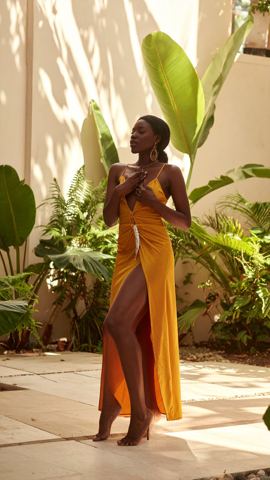 A woman in a saffron dress stands among broad leaves, a hand on her heart, looking away into the light.
