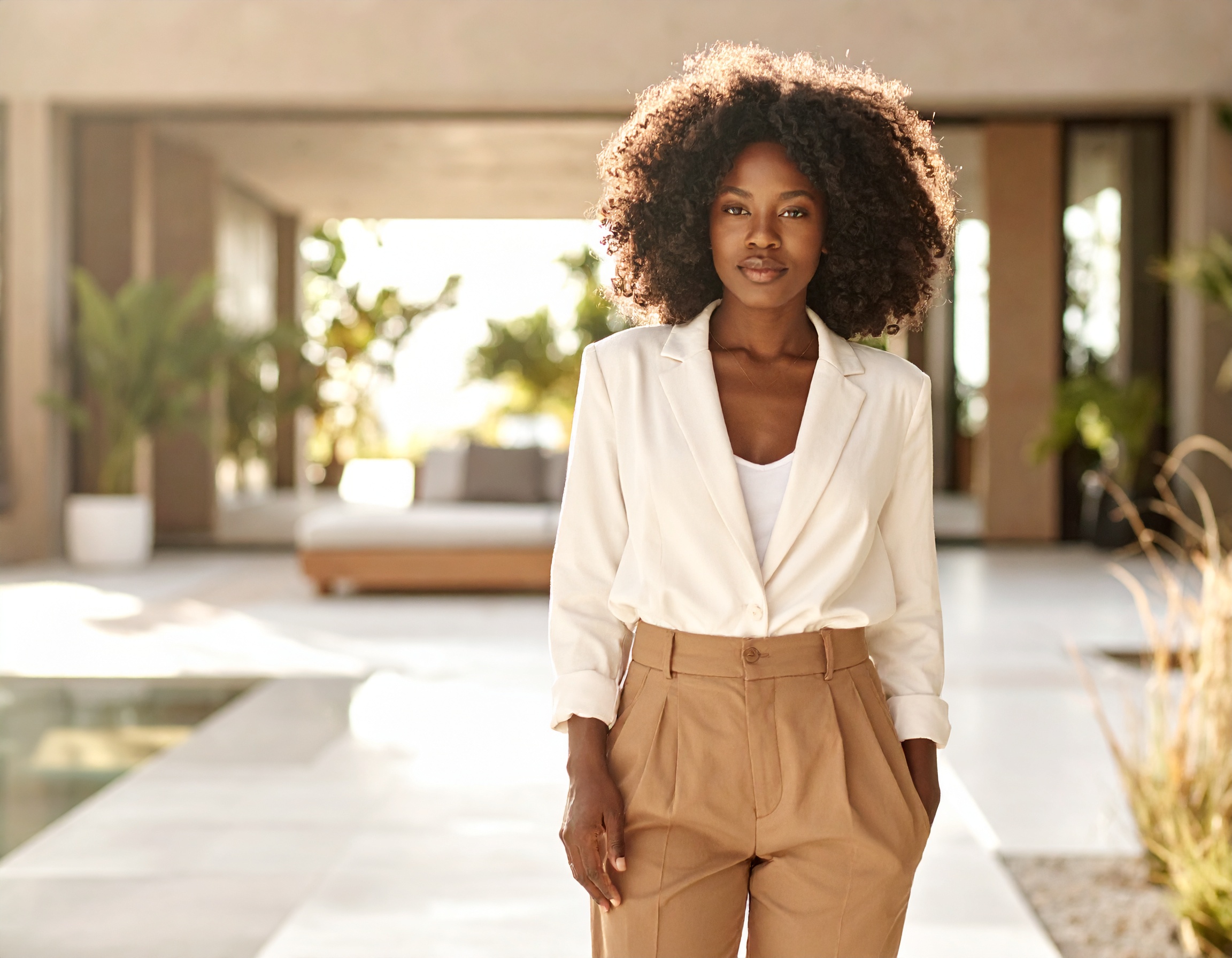 A woman in a white blazer and camel trousers, still, in a bright courtyard