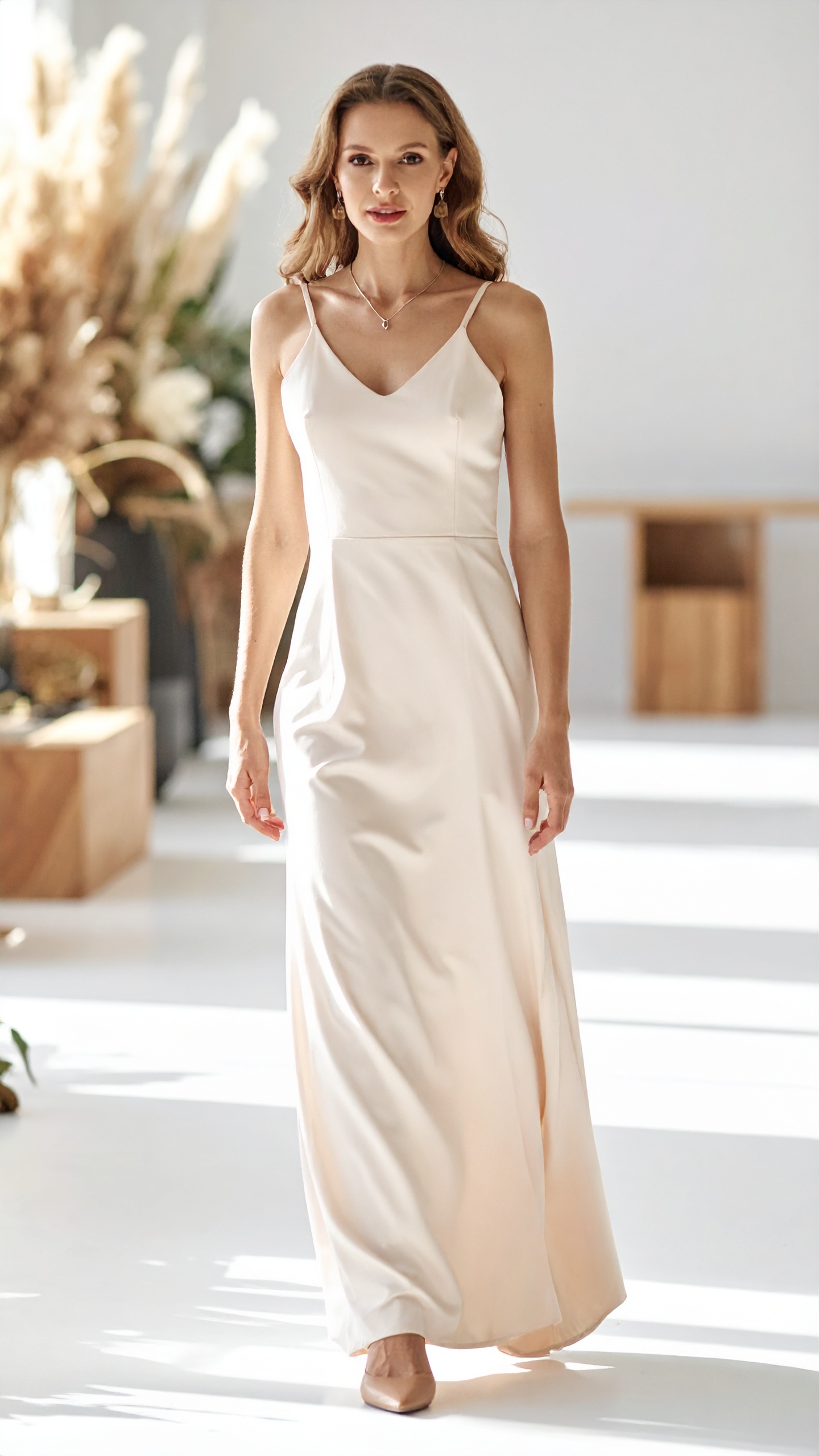 A woman in a slip gown walking a bright hall, pampas and low daylight behind her