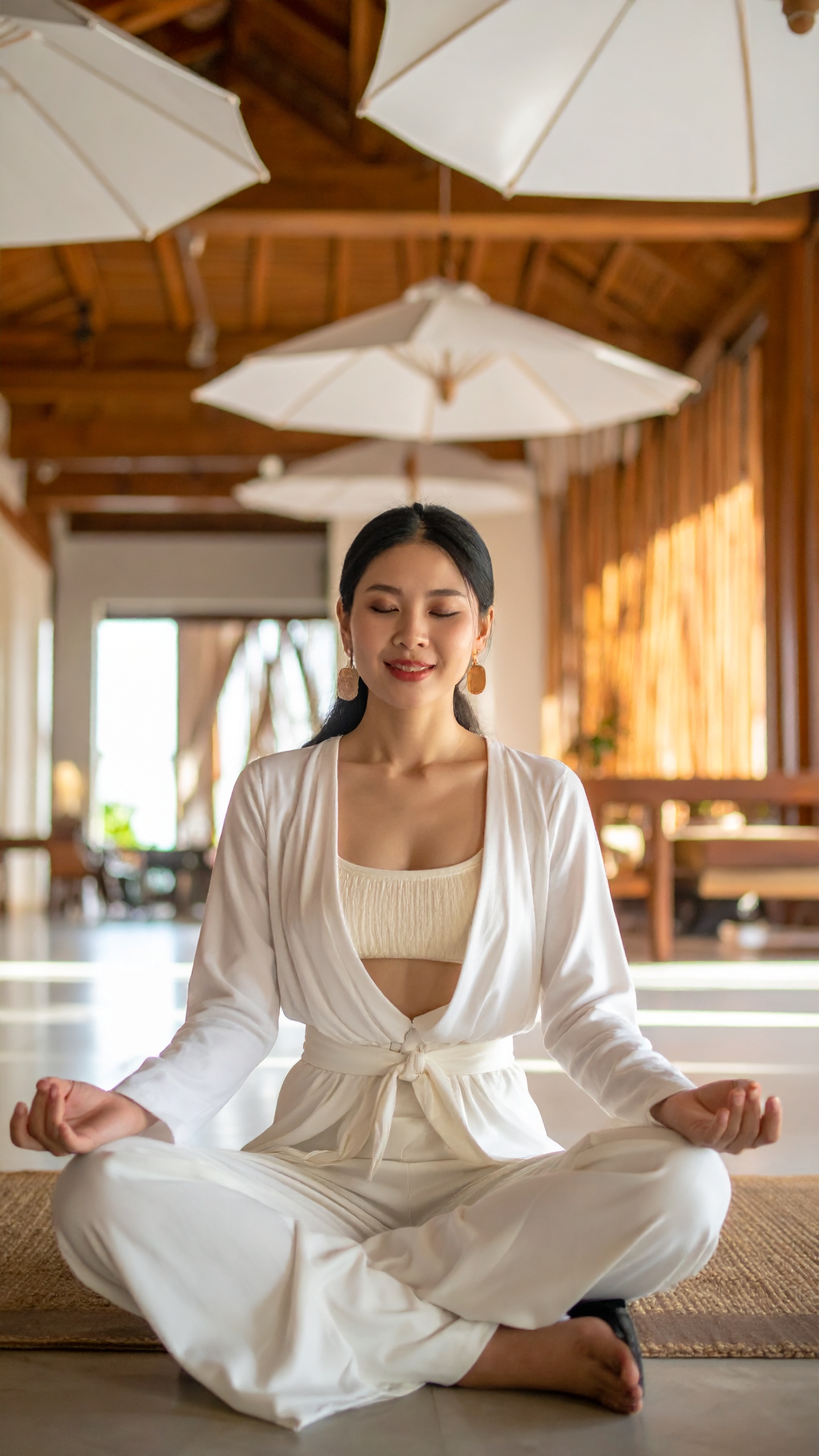 A woman seated in lotus on a wooden floor in a wide sunlit room, garden beyond the glass